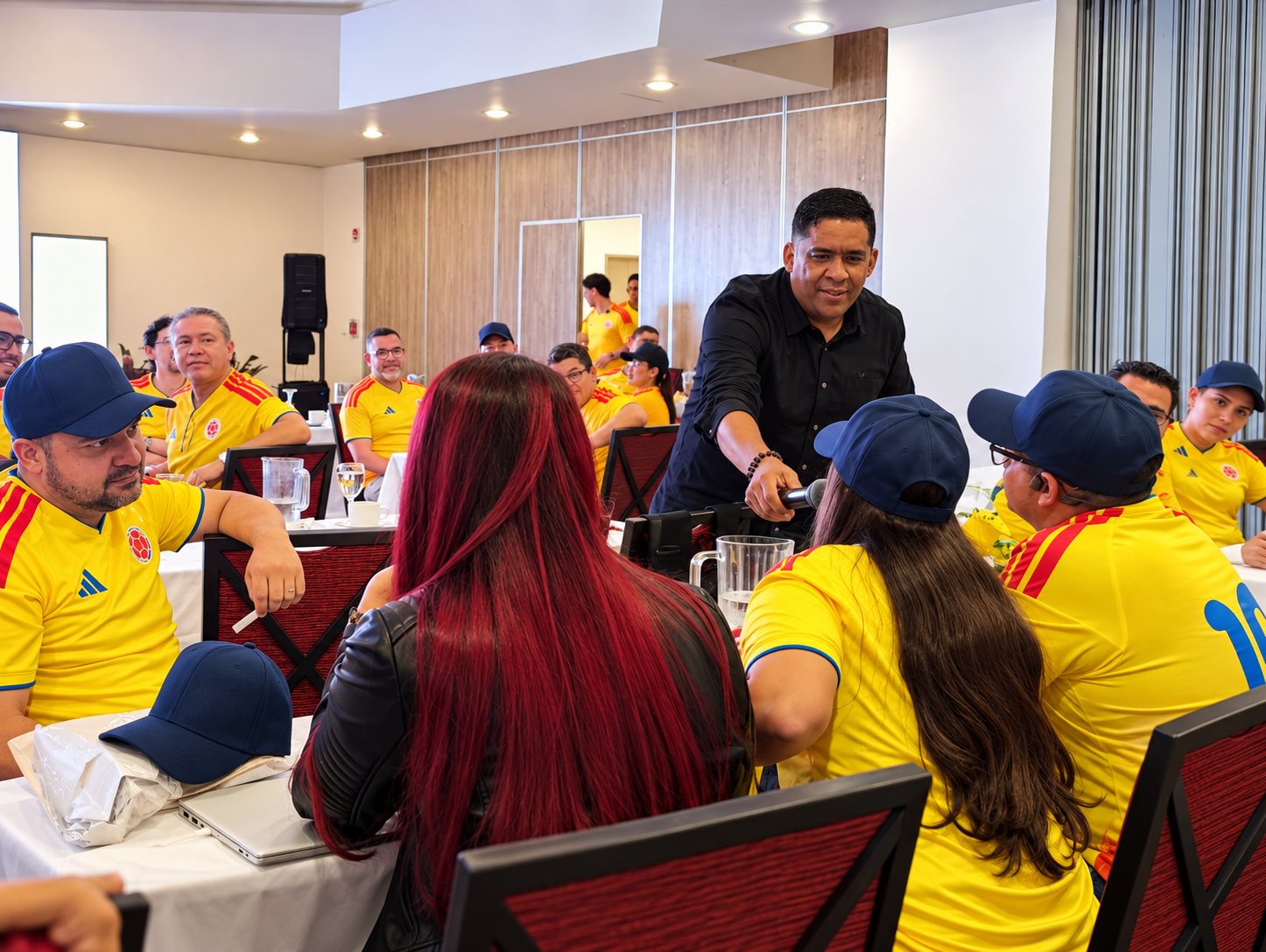 El facilitador entregando el micrófono a una participante durante el trabajo en mesas, con el grupo en camisetas amarillas